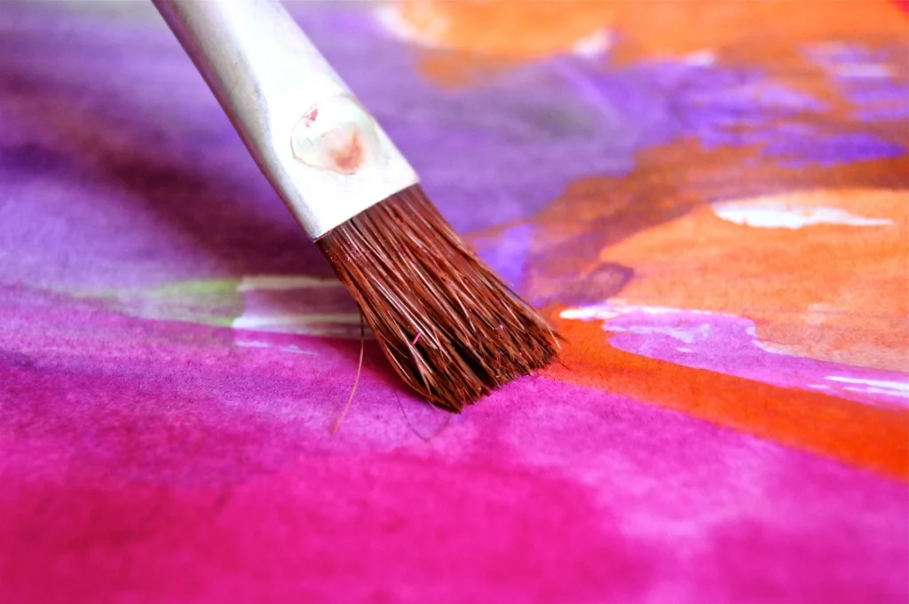 A close-up of a paintbrush applying vibrant wall paint colors, including shades of purple, orange, and pink, on a canvas. The image captures the detailed texture of the brush bristles and the smooth blending of the colors, showcasing the artistry and precision in wall painting.