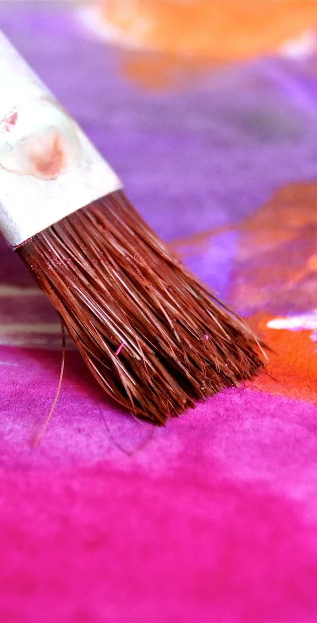 A close-up of a paintbrush applying vibrant wall paint colors, including shades of purple, orange, and pink, on a canvas. The image captures the detailed texture of the brush bristles and the smooth blending of the colors, showcasing the artistry and precision in wall painting.