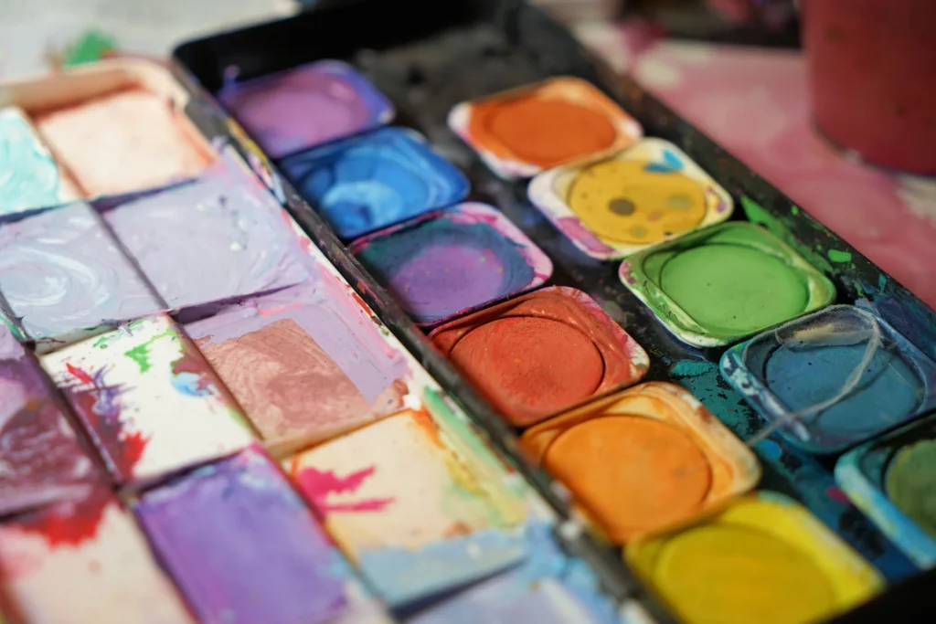 A close-up view of an artist's palette filled with various vibrant colors of paint, including shades of green, blue, orange, yellow, purple, and pink. The paint is organized in small, square pans, each showing signs of use with visible brush strokes and mixing. This image highlights the diverse color options available for wall paint, emphasizing creativity and the importance of color in interior design.